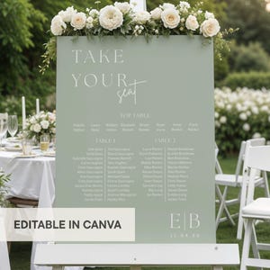May include: A sage green wedding seating chart with white floral accents. The sign features the words "TAKE YOUR SEAT" and guest names organized by table. The bottom of the sign reads "EDITABLE IN CANVA".