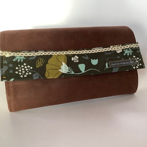 Könnte beinhalten: Eine braune Leder-Clutch mit einem floralen Stoffklappen. Die Klappe hat einen weißen Spitzenbesatz und ein kleines Etikett mit der Aufschrift "mme-rose.de".