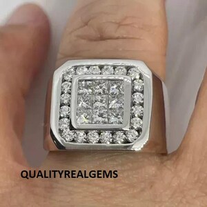 May include: A silver ring with a square-shaped center stone surrounded by smaller stones. The ring has a thick band.