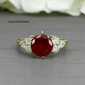 Könnte beinhalten: Ein goldener Ring mit einem großen, runden, roten Rubin-Edelstein, der von kleineren, runden, weißen Diamanten umgeben ist.