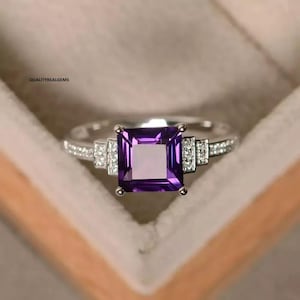 May include: A silver ring with a square-shaped purple amethyst gemstone surrounded by smaller clear gemstones.