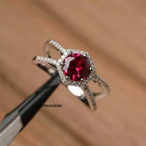Può includere: Un anello d'argento con una grande gemma rosso scuro rotonda al centro, circondata da pietre chiare più piccole. L'anello ha un design a gambo diviso ed è posto su uno sfondo di legno sfocato. Il testo "QUALITYREALGEMS" è visibile.