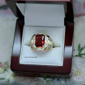 May include: A gold ring with a large, rectangular, red gemstone at the center. The ring is set in a gold band with smaller, clear gemstones and decorative detailing. The ring is displayed in a dark red jewelry box.
