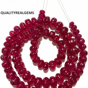 Puede incluir: Hebras de cuentas de piedras preciosas de color rojo intenso. Las cuentas tienen un diseño tallado y acanalado y están ensartadas en un cordón fino y transparente. La imagen incluye el texto "QUALITYREALGEMS". Es probable que estas cuentas se utilicen para la fabricación de joyas.