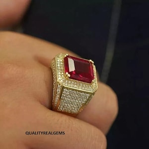 May include: A gold ring with a large, red, emerald-cut gemstone surrounded by smaller, clear gemstones.