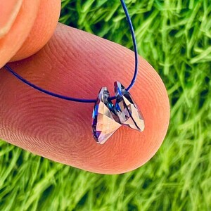 May include: A pair of iridescent crystal pendants, each with multiple facets, are strung on a thin blue cord. The crystals reflect a range of colors, including blue, purple, and silver. The background is a blurred green.