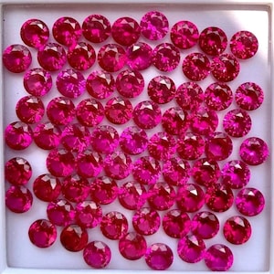May include: A collection of round, faceted, hot pink gemstones. The gems are arranged closely together, filling a white square container. The stones have a brilliant cut, reflecting light and creating a sparkling effect. The overall impression is one of luxury and value.