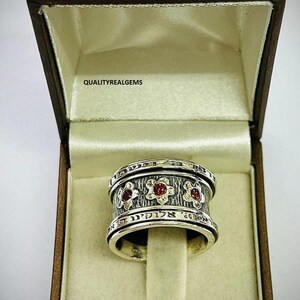 May include: A silver ring with three red gemstones set in a floral design. The ring is inscribed with Hebrew text and is displayed in a brown box with the text "QUALITYREALGEMS".