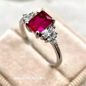 Könnte beinhalten: Ein silberner Ring mit einem großen, smaragdgeschliffenen Rubin und kleineren, klaren Edelsteinen, die darum herumgesetzt sind.