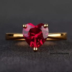 May include: A gold ring featuring a heart-shaped, deep red gemstone. The gemstone is held in place by four gold prongs. The ring band is a simple, polished gold. The background is a dark gray.