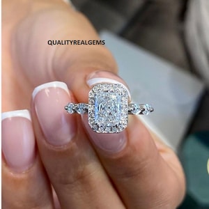 May include: A silver ring with a large, rectangular, clear diamond surrounded by smaller diamonds. The diamond is set in a halo setting. The ring is on a finger with white nail polish.