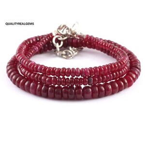 Op de afbeelding: Een ketting bestaande uit drie strengen dieprode robijn kralen. De ronde kralen zijn verbonden met een zilverkleurige sluiting. Het sieraad wordt gepresenteerd tegen een witte achtergrond. Een elegant accessoire.