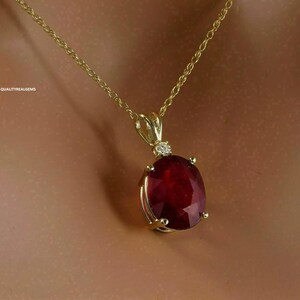 May include: A gold necklace with a large, oval-cut, deep red ruby pendant. A small diamond accents the top of the pendant. The necklace is displayed against a neutral background, highlighting the jewelry.