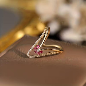 May include: A gold-colored ring with a floral design. The ring features a small, red flower with a white center stone. The band is adorned with small, clear stones. The ring is set on a brown surface.