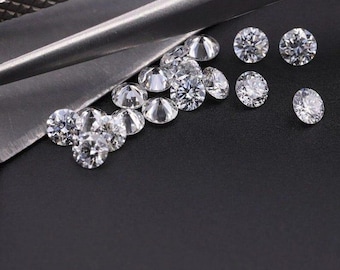 Może przedstawiać: Kolekcja błyszczących, okrągłych diamentów ułożonych na ciemnej powierzchni. Diamenty są przejrzyste i odbijają światło, ukazując swoje fasetki. Częściowo widoczne jest srebrne narzędzie, sugerujące warsztat jubilera.