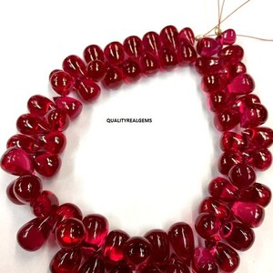 May include: A strand of deep red ruby gemstone beads in a teardrop shape. The beads are strung on a thin wire and are arranged in a circular pattern. The beads are all approximately the same size and shape. The image is taken on a white background.