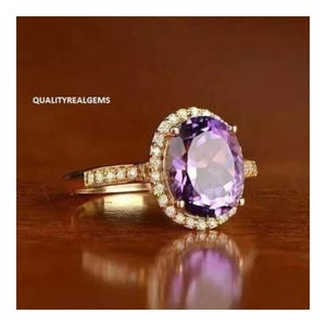 May include: A gold ring with a large oval amethyst gemstone surrounded by a halo of smaller diamonds. The ring is set on a wooden surface.
