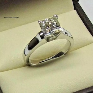 May include: A silver engagement ring with a square-shaped diamond in the center. The ring is set in a simple band.