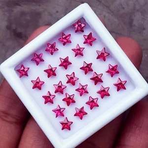 Peut inclure: Un plateau carré blanc rempli de petits gemmes en forme d'étoile, de couleur rouge rubis. Les pierres précieuses sont disposées en rangées et en colonnes, créant un motif symétrique. Le plateau est tenu dans une main.
