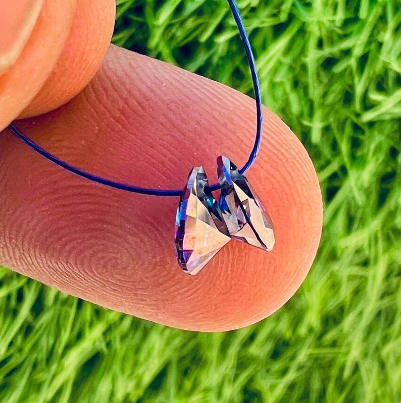 May include: A pair of iridescent crystal pendants, each with multiple facets, are strung on a thin blue cord. The crystals reflect a range of colors, including blue, purple, and silver. The background is a blurred green.