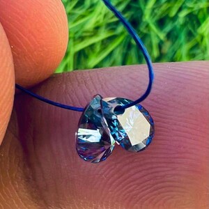 May include: Two faceted, teardrop-shaped crystals with a blue and purple iridescent sheen are strung on a thin blue cord. The crystals are held between fingers, with a blurred green background.