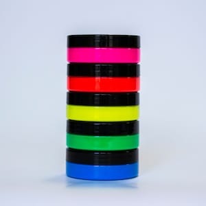 May include: A stack of six small round containers with black lids. The containers are filled with bright neon colors: pink, red, yellow, green, and blue.