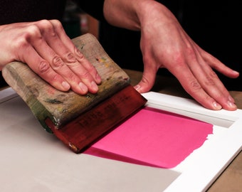 Può includere: Una persona utilizza una spatola di legno per applicare inchiostro rosa su un telaio per serigrafia.