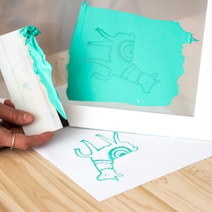 May include: A teal llama design is printed on a white sheet of paper using a screen printing frame. The frame is made of white wood and has a clear plastic screen. A teal squeegee is used to apply the ink to the screen.