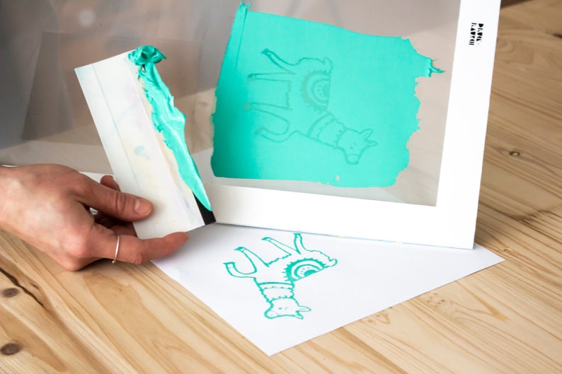 May include: A teal llama design is printed on a white sheet of paper using a screen printing frame. The frame is made of white wood and has a clear plastic screen. A teal squeegee is used to apply the ink to the screen.