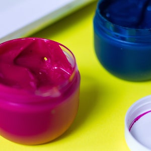 Pu&ograve; includere: Due piccoli barattoli di vernice, uno rosa e uno blu, su una superficie gialla. La vernice rosa &egrave; in un barattolo trasparente e la vernice blu &egrave; in un barattolo bianco.