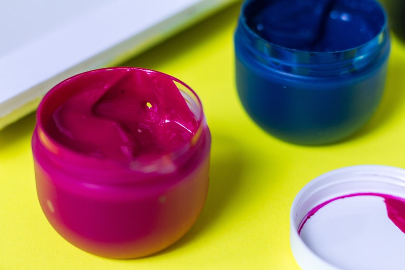 Pu&ograve; includere: Due piccoli barattoli di vernice, uno rosa e uno blu, su una superficie gialla. La vernice rosa &egrave; in un barattolo trasparente e la vernice blu &egrave; in un barattolo bianco.