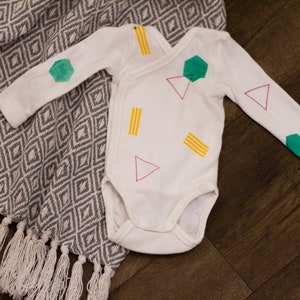Pu&ograve; includere: Body bianco per neonato con un motivo geometrico in verde, giallo e rosa. Il motivo comprende triangoli, esagoni e rettangoli.