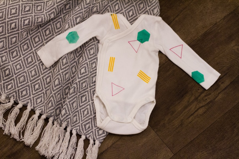 Pu&ograve; includere: Body bianco per neonato con un motivo geometrico in verde, giallo e rosa. Il motivo comprende triangoli, esagoni e rettangoli.