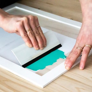 May include: A person using a white and black squeegee to smooth out a turquoise blue vinyl sticker on a white frame.