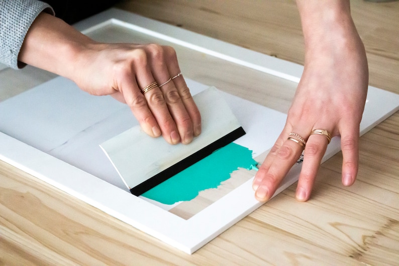May include: A person using a white and black squeegee to smooth out a turquoise blue vinyl sticker on a white frame.