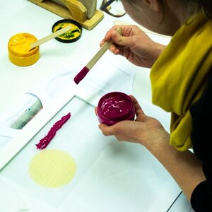 May include: A person is using a wooden stick to apply pink paint to a white t-shirt. There are two containers of paint, one yellow and one pink.