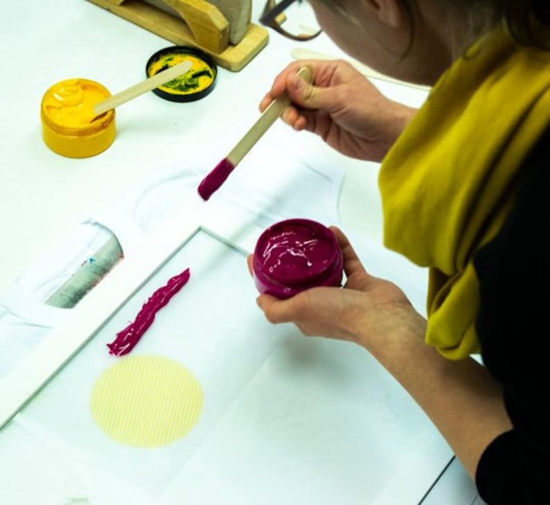 May include: A person is using a wooden stick to apply pink paint to a white t-shirt. There are two containers of paint, one yellow and one pink.