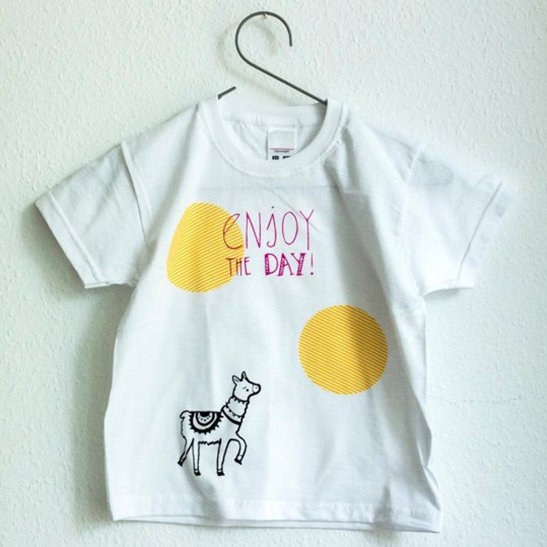 May include: White t-shirt with a yellow sun, a llama, and the text "Enjoy the day!" in pink.
