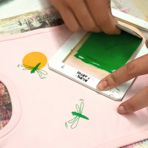 Pu&ograve; includere: Una persona utilizza un telaio per serigrafia per applicare vernice verde su un tessuto rosa con un motivo a libellula. Il telaio ha il testo "DRINK" e "MADUI" su di esso.