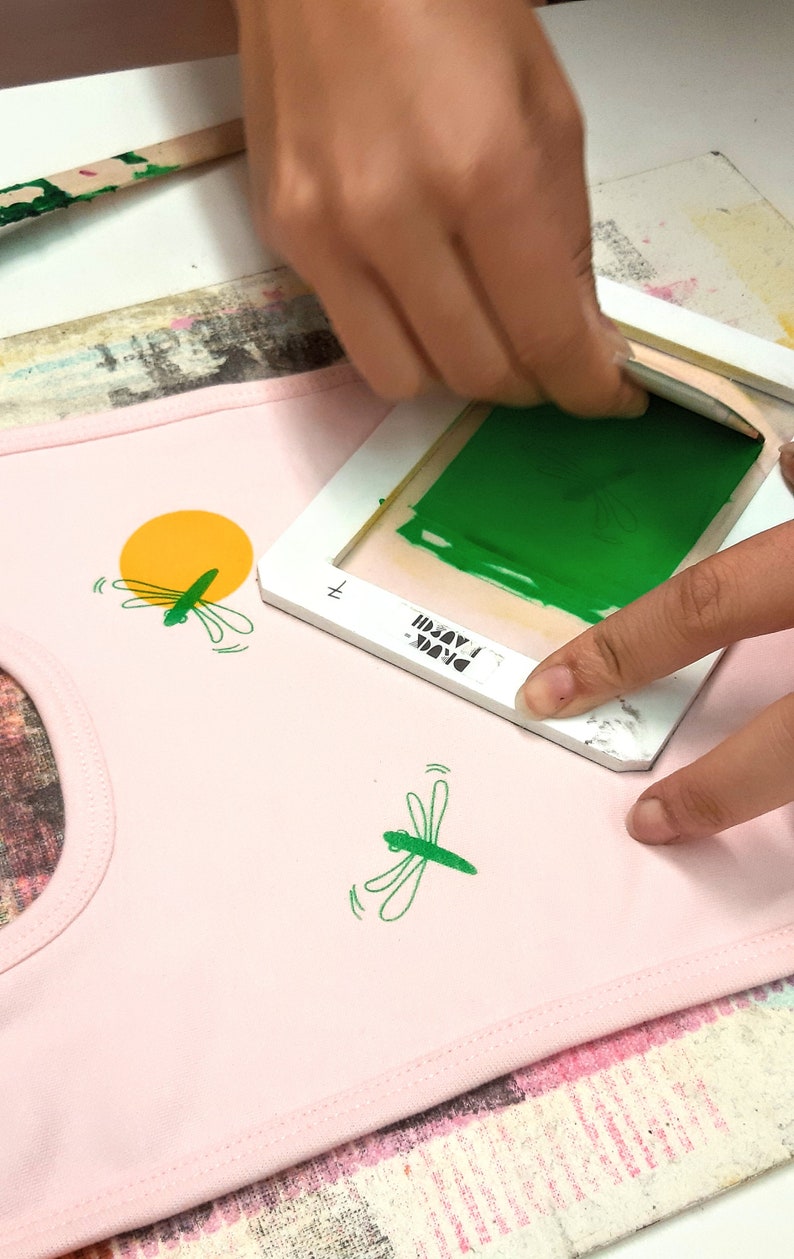 Pu&ograve; includere: Una persona utilizza un telaio per serigrafia per applicare vernice verde su un tessuto rosa con un motivo a libellula. Il telaio ha il testo "DRINK" e "MADUI" su di esso.