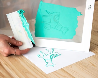 May include: A teal llama design is printed on a white sheet of paper using a screen printing frame. The frame is made of white wood and has a clear plastic screen. A teal squeegee is used to apply the ink to the screen.