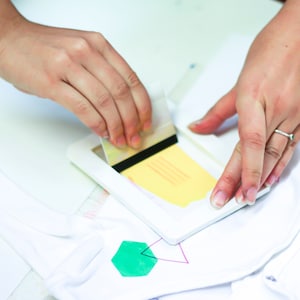 Pu&ograve; includere: Una persona utilizza una spatola per applicare un disegno giallo su un capo di abbigliamento bianco. Il disegno viene applicato tramite uno stencil.