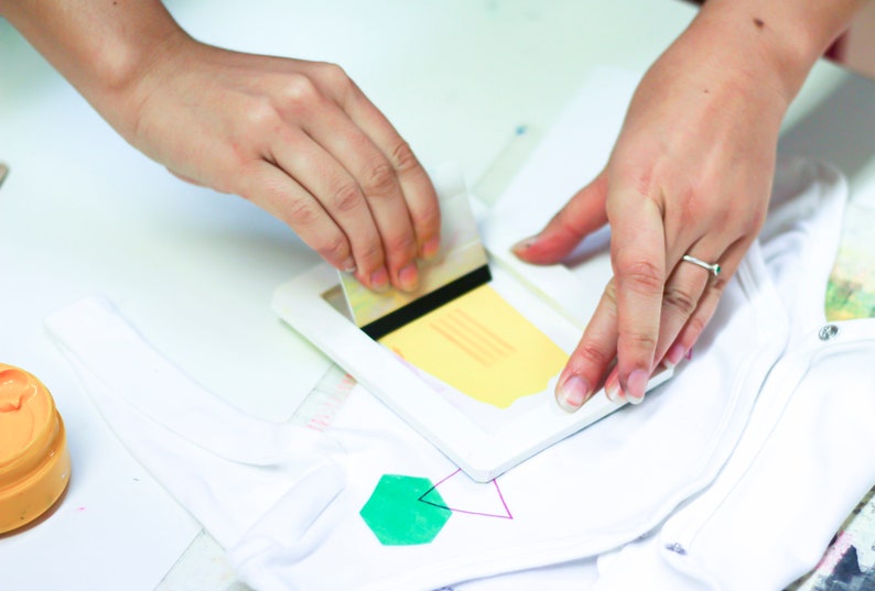 Pu&ograve; includere: Una persona utilizza una spatola per applicare un disegno giallo su un capo di abbigliamento bianco. Il disegno viene applicato tramite uno stencil.