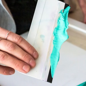 May include: A white plastic squeegee with a black edge, being used to spread a bright turquoise blue paint or ink.