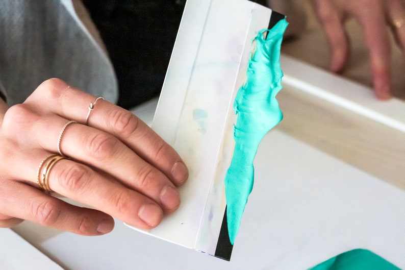 May include: A white plastic squeegee with a black edge, being used to spread a bright turquoise blue paint or ink.