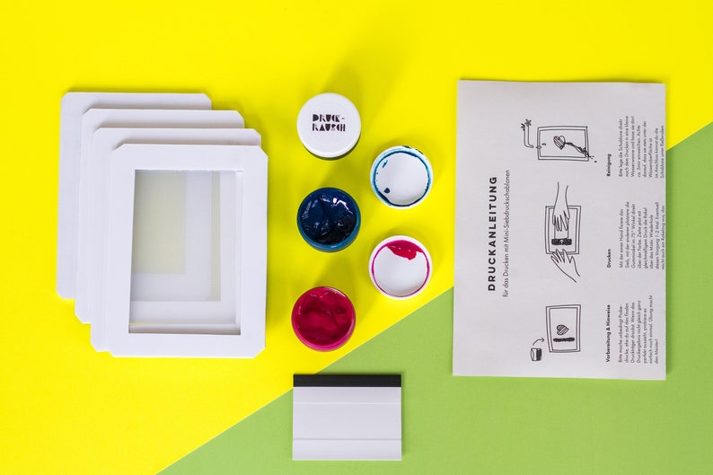 Pu&ograve; includere: Un kit di serigrafia con quattro telai bianchi, quattro barattoli di vernice blu, rosa e bianca, una spatola e istruzioni in tedesco. Il kit &egrave; su una superficie gialla e verde.