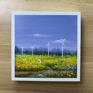 May include: A painting of a field of yellow flowers with a single figure in a white dress and blue hat standing in the middle of the field. There are four wind turbines in the background against a blue sky.