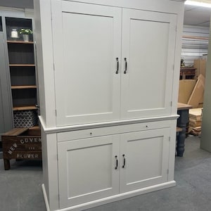 May include: A white cabinet with two large doors on top and two smaller doors on the bottom. The cabinet has black metal handles. The cabinet is in a room with a wooden floor.