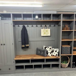 May include: A grey wooden hallway storage unit with a bench, coat hooks, and shelves. Includes a cabinet, open shelves, and a bench with shoe storage. Features a tote bag, cushions, and decorative items.