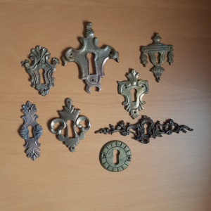 Peut inclure: Une collection de huit écussons de serrure en laiton antique avec des motifs complexes. Les écussons sont disposés sur une surface en bois brun clair.